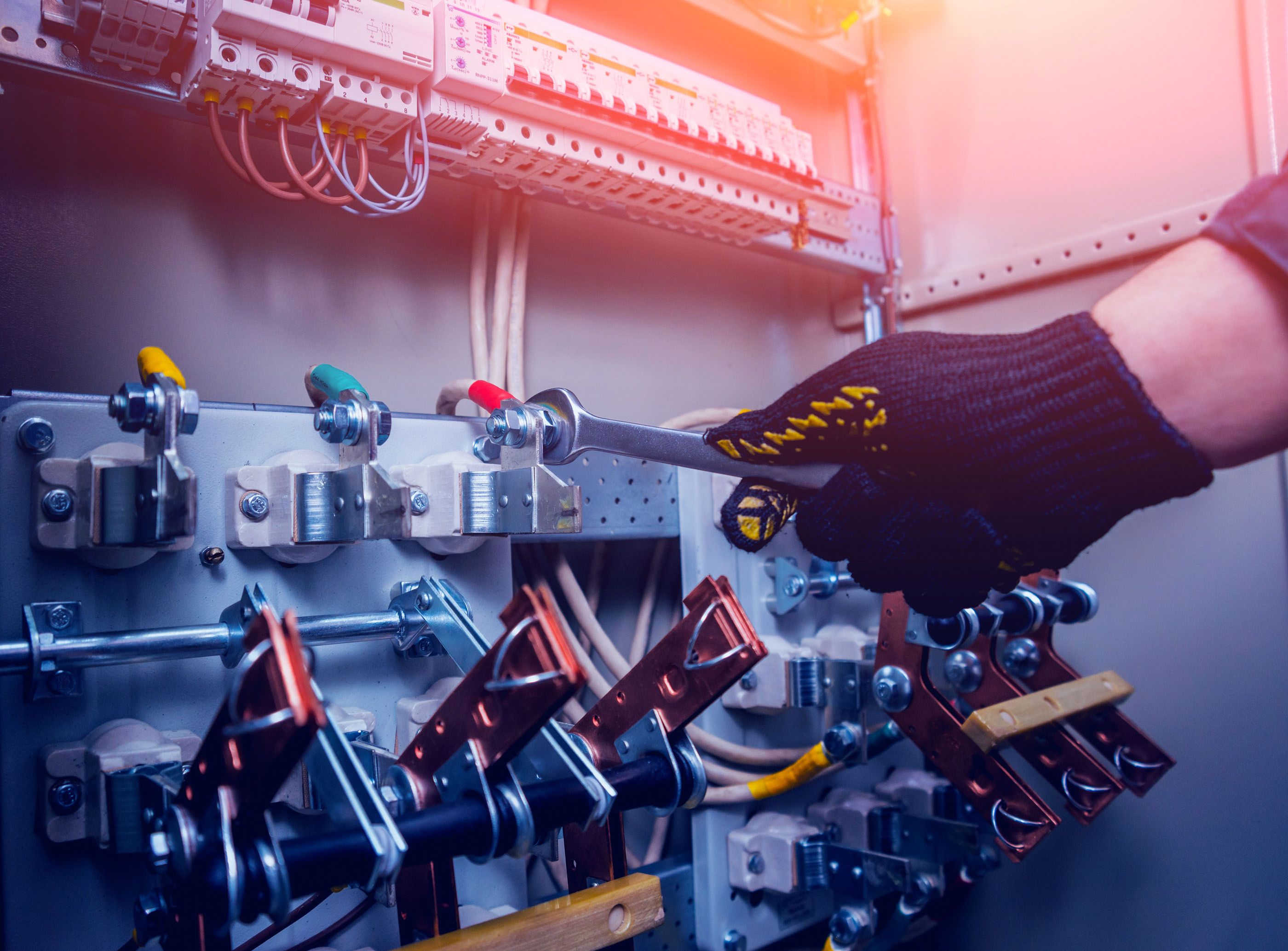 Eine Hand in einem schwarzen Handschuh hält einen Schraubenschlüssel und greift nach einem Schalter in einem elektrischen Schaltschrank. Sichtbare Komponenten sind Schalter, Kabel und Verbindungselemente.